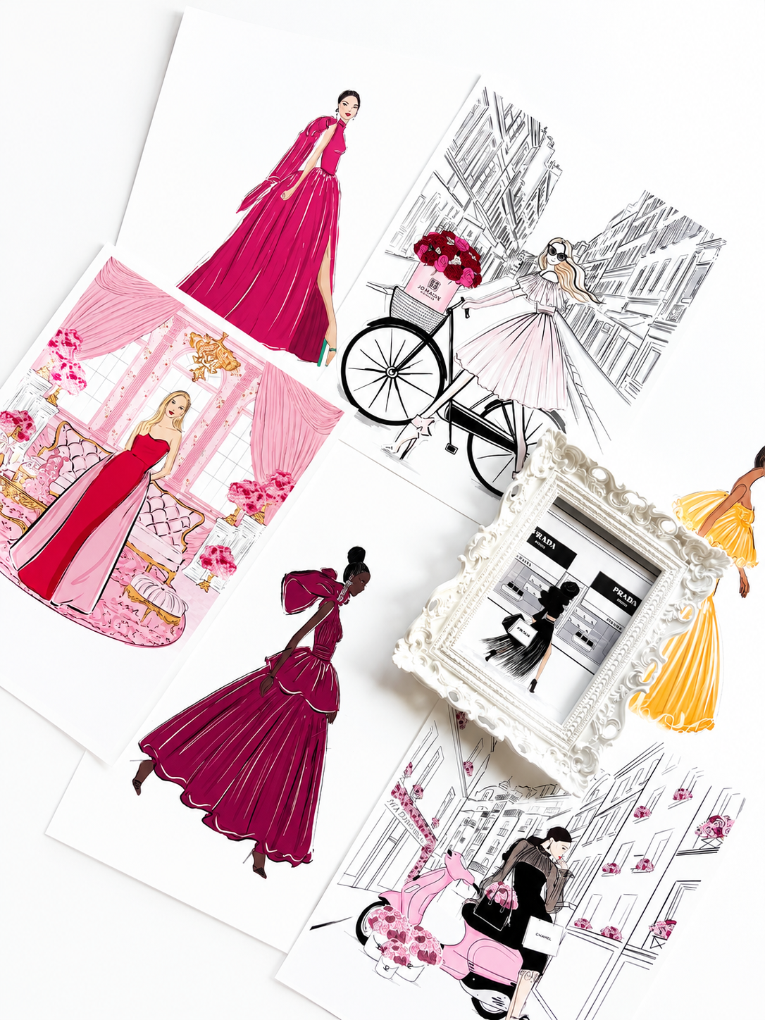 A styled flat-lay of fashion illustration art prints featuring elegant women in pink and magenta gowns, a bicycle with a flower basket, and a small ornate white picture frame on a white background.